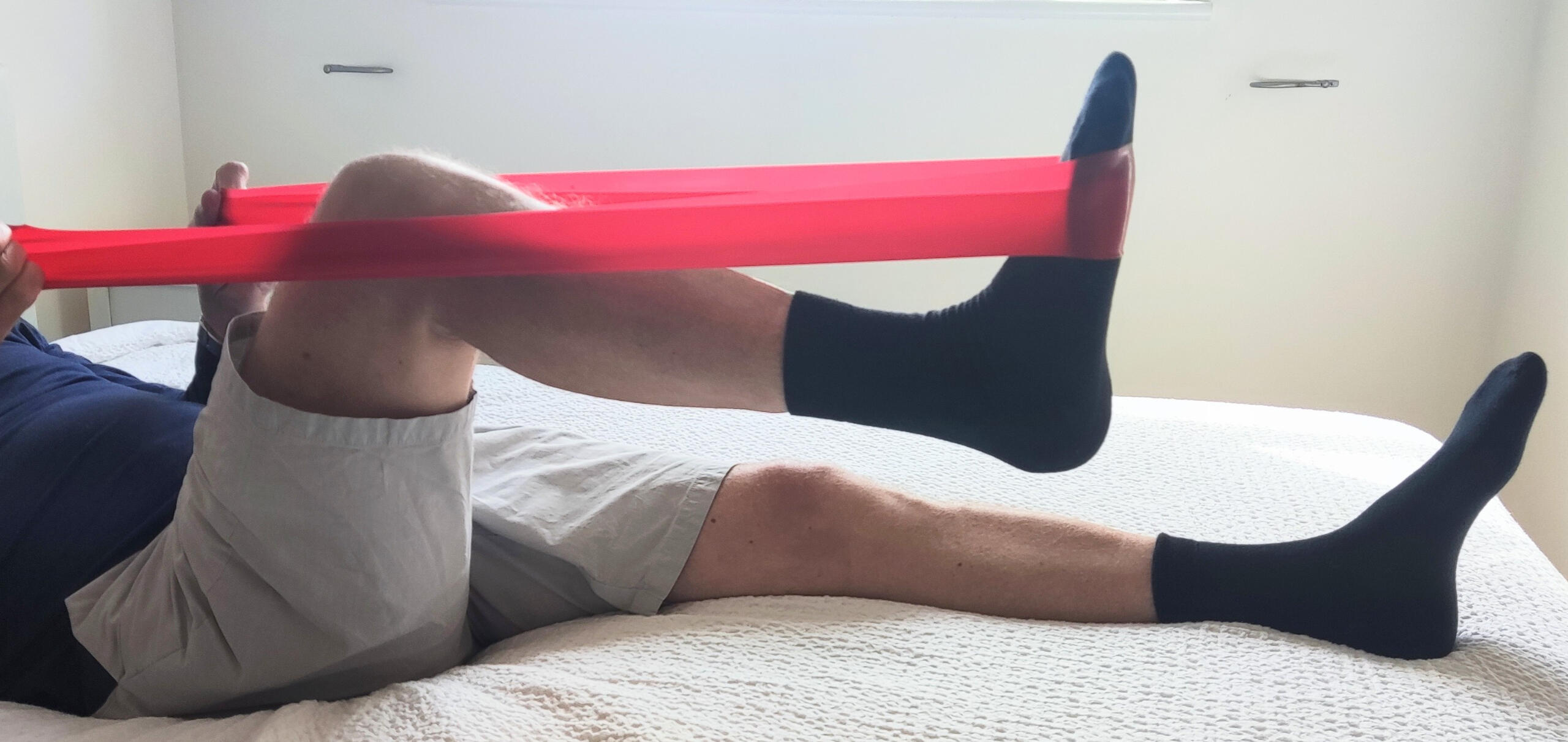 Man using a therapy band Person lying on a bed performing a leg exercise using a red resistance band, stretching one leg while keeping the other flat